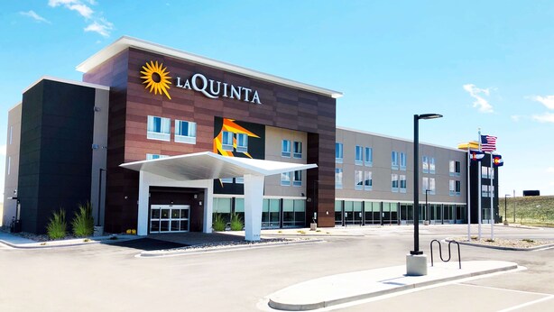 Un hotel La Quinta con un diseño moderno, que presenta un prominente logo de sol, una entrada cubierta y un estacionamiento.