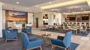 Un moderno vestíbulo de hotel con sillones tapizados en azul, un mostrador de recepción, un área de bar y un gran mural con un paisaje de atardecer.