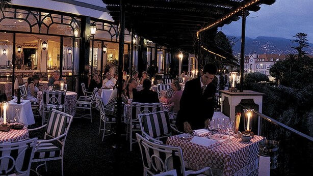 An outdoor dining area with tables and chairs, overlooking a coastal landscape with buildings and a view of the sea.