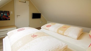 Une chambre avec deux lits, chacun recouvert d’une couverture blanche ornée d’un motif brodé rouge. Un téléviseur est fixé au mur, au-dessus des lits.