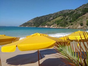 A beach with yellow umbrellas and a clear view of the sea and a hillside in the background. - Orlanova Hotel (Arraial do Cabo)