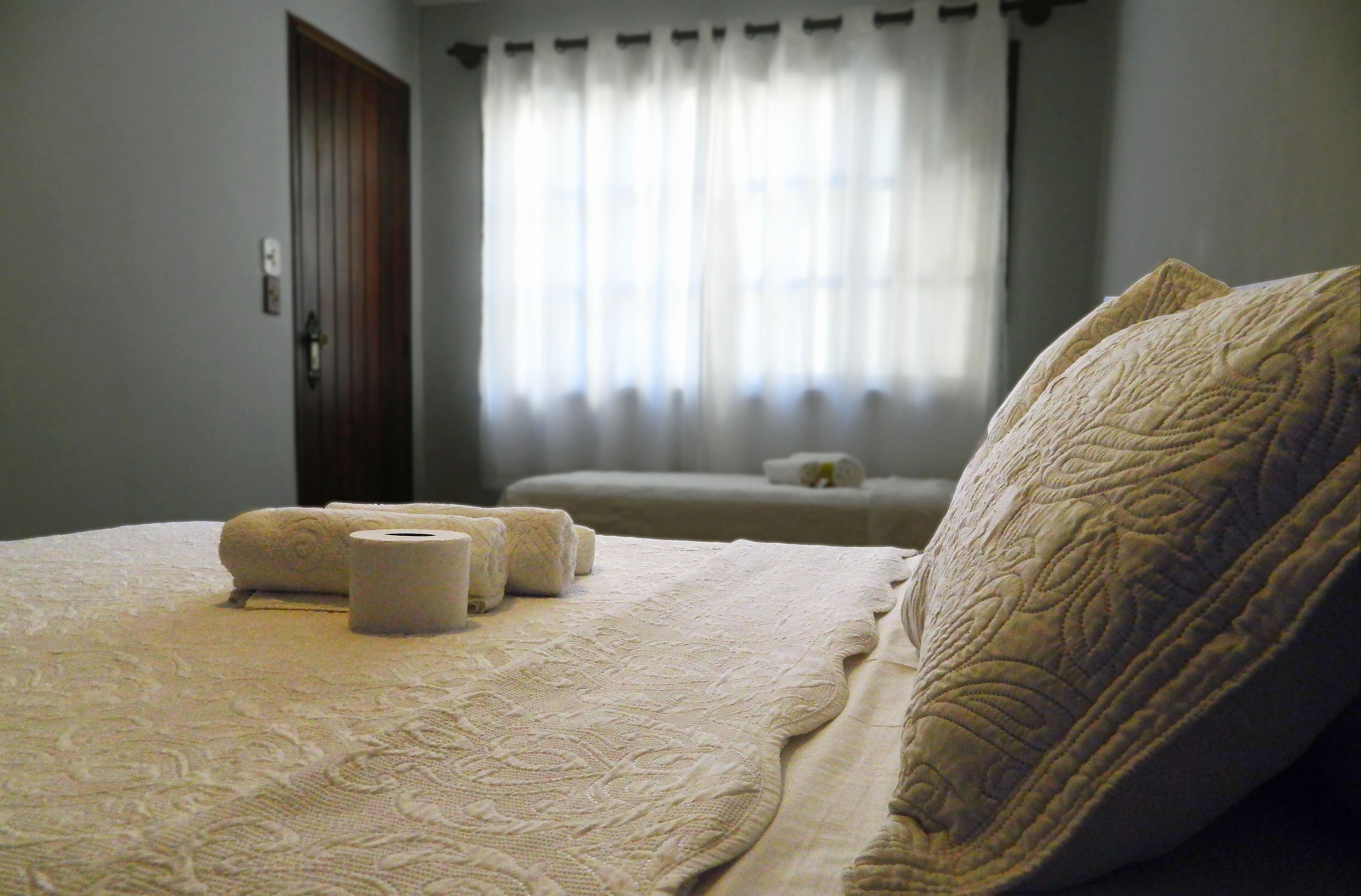 A neatly made bed with a patterned bedspread and pillows, a bedside table with a lamp, and a window with sheer curtains.