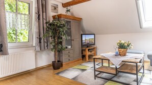 Une pièce chaleureuse avec un plafond en bois, un radiateur, un téléviseur posé sur un meuble en bois, une table recouverte d’une nappe, une plante en pot et une fenêtre avec des rideaux.