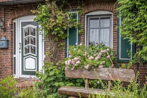 Ein Backsteinhaus mit grüner Tür und Fensterläden, einer Holzbank und rosa Blumen.