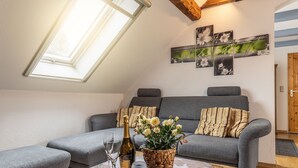Un salon chaleureux avec un canapé gris, une petite table sur laquelle se trouvent une bouteille et des verres, et une fenêtre donnant sur un paysage verdoyant.