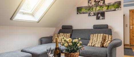 Uma sala de estar acolhedora, com um sofá cinzento, uma mesinha com uma garrafa e copos, e uma janela com vista para o verde.