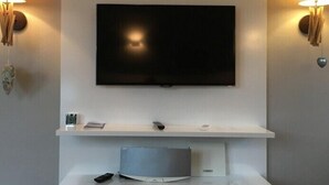 Ein modernes Wohnzimmer mit einem Flachbildfernseher an einer weißen Wand, der von zwei Wandlampen mit dekorativen Elementen eingerahmt wird. Darunter befindet sich eine weiße Ablage mit einer Fernbedienung und einem kleinen Lautsprecher.