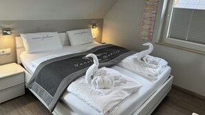 Ein ordentlich bezogenes Bett mit weißen Leinen, zwei Kissen mit Schriftzug, einer grauen Decke und zwei zu Schwänen geformten Handtüchern.