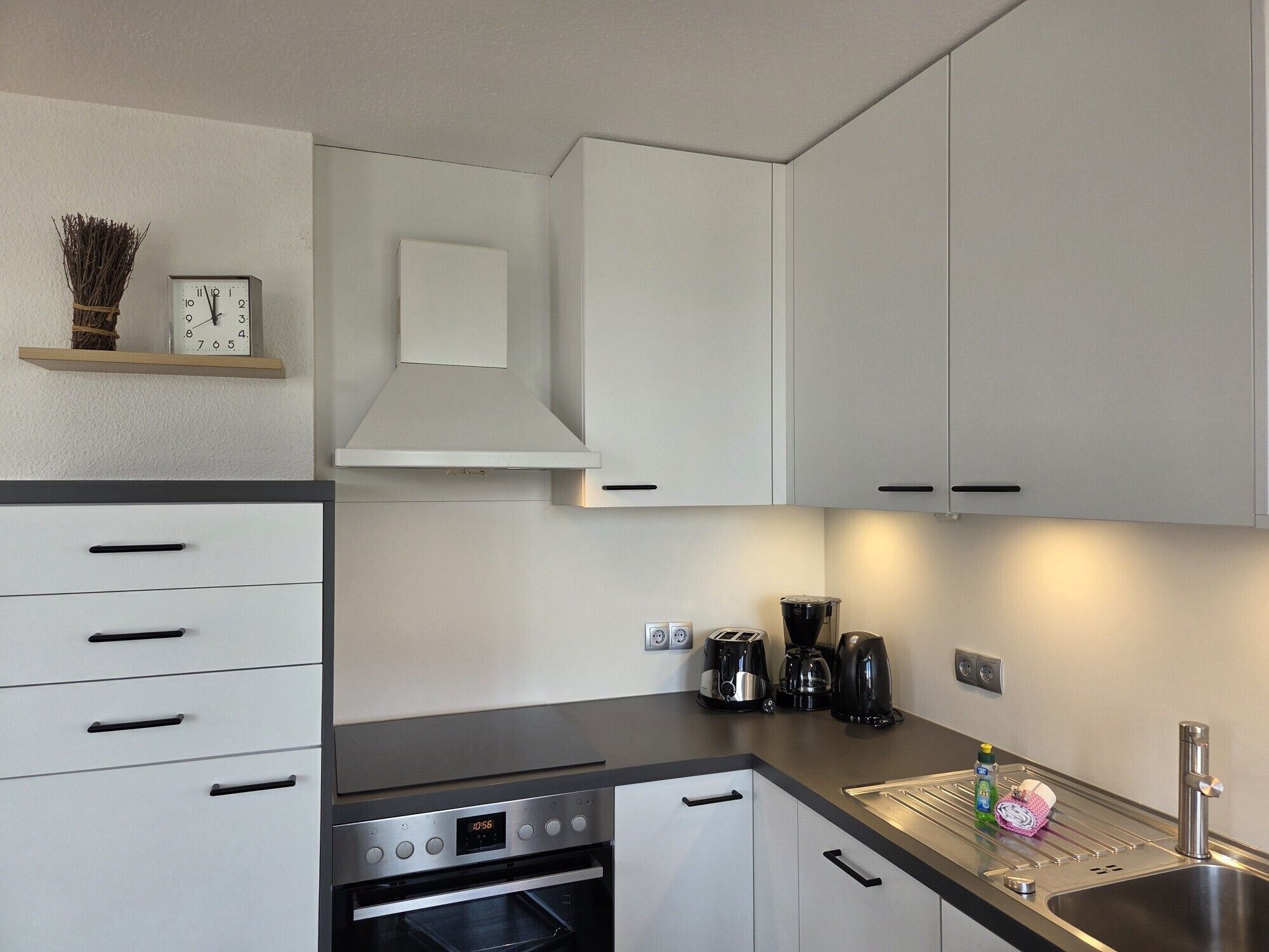 Een moderne keuken met witte kastjes, een inbouwoven, een koffiezetapparaat en een klok aan de muur.