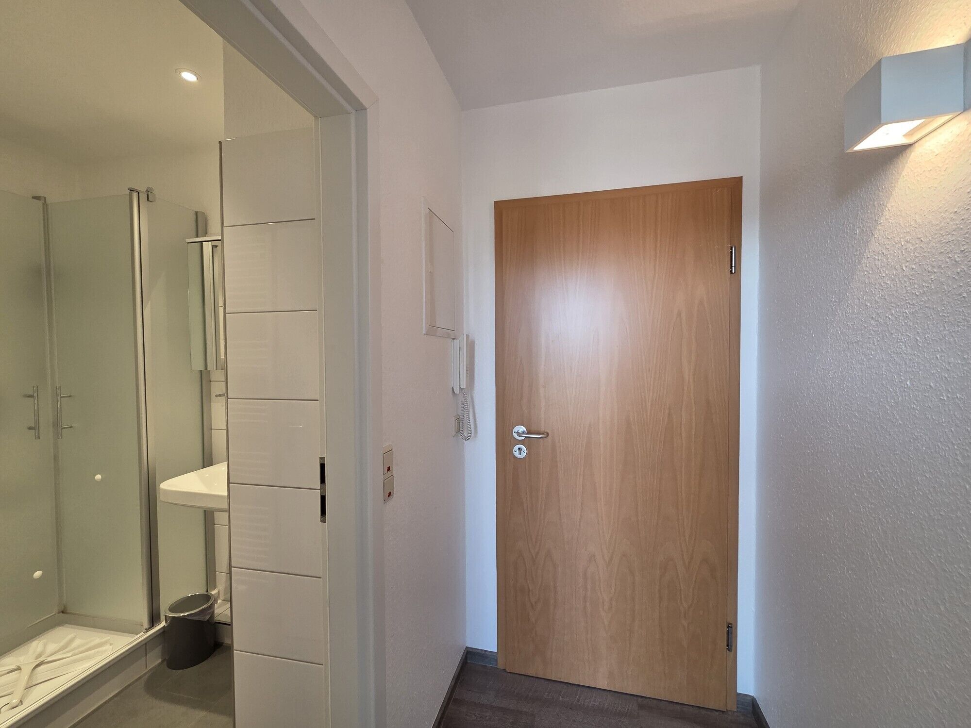 A hallway with a wooden door, a glass shower enclosure, and a white tiled wall.
