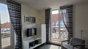 A room with a TV on a white stand, a grey chair, and a window with curtains. - Duhner Düne apartment 25, WLAN, elevator, roof terrace, close to the beach, non-smoking (Cuxhaven)