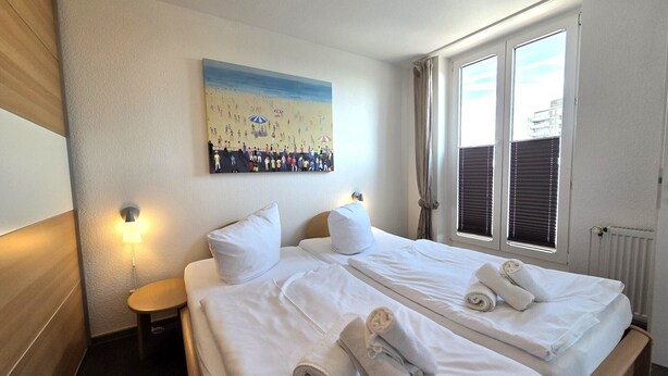A double bed with white linens, a painting of a beach scene, and a window with blinds. - Duhner Düne apartment 25, WLAN, elevator, roof terrace, close to the beach, non-smoking (Cuxhaven)