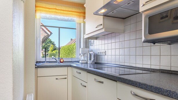 Una cocina moderna con armarios blancos, encimera de granito y ventana con vistas a una casa y follaje.