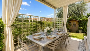 Un área de comedor al aire libre con una mesa preparada para cuatro personas, rodeada de sillas, bajo un porche cubierto.