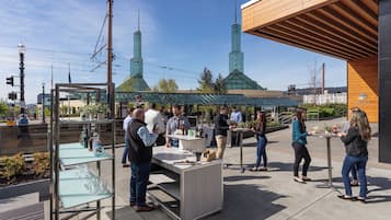 Evento social al aire libre con zona de bar, gente interactuando y un edificio moderno con estructuras de vidrio al fondo.