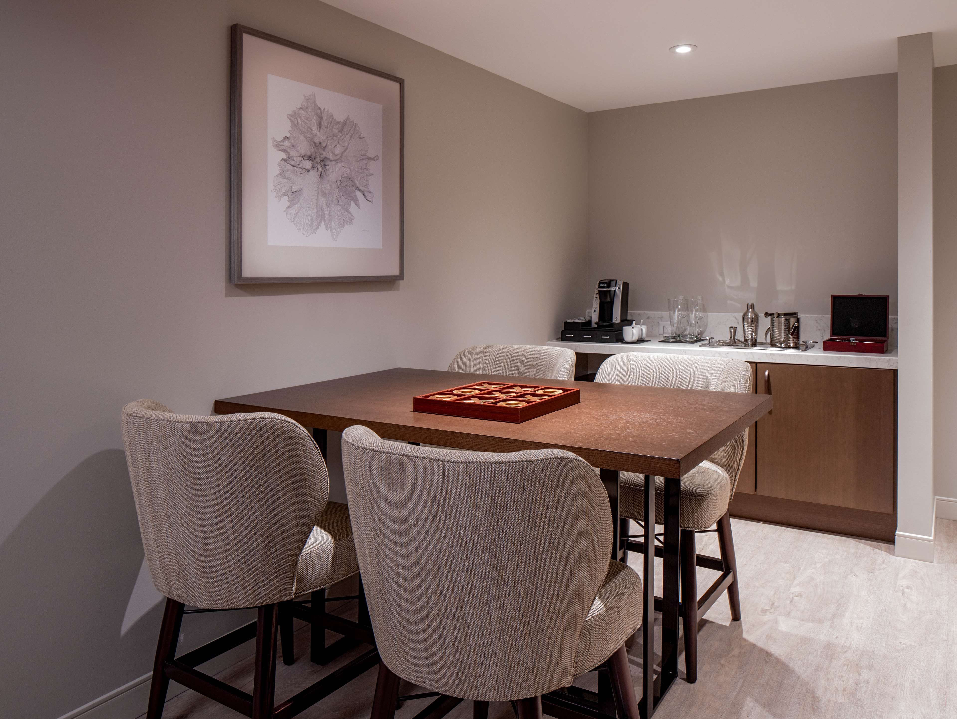 A dining area with a wooden table and four chairs, a framed artwork on the wall, and a kitchen area with appliances and glassware.