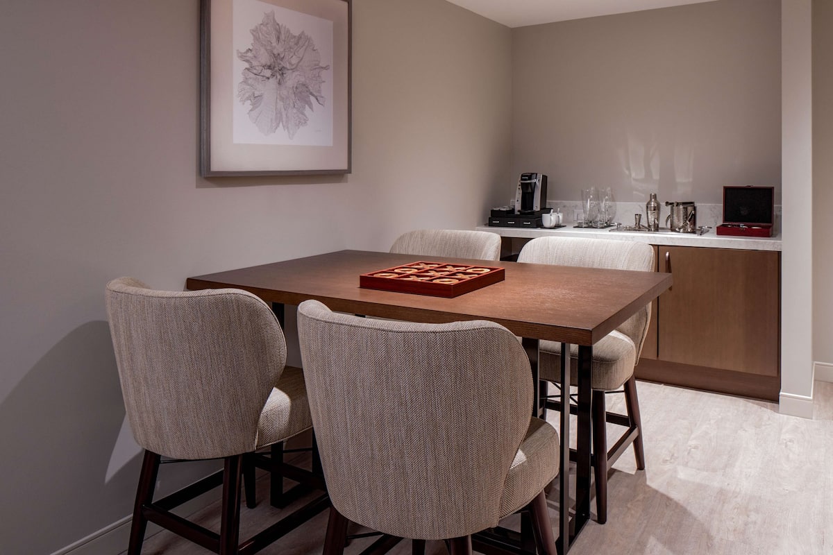 A dining area with a wooden table and four chairs, a framed artwork on the wall, and a kitchen area with appliances and glassware.