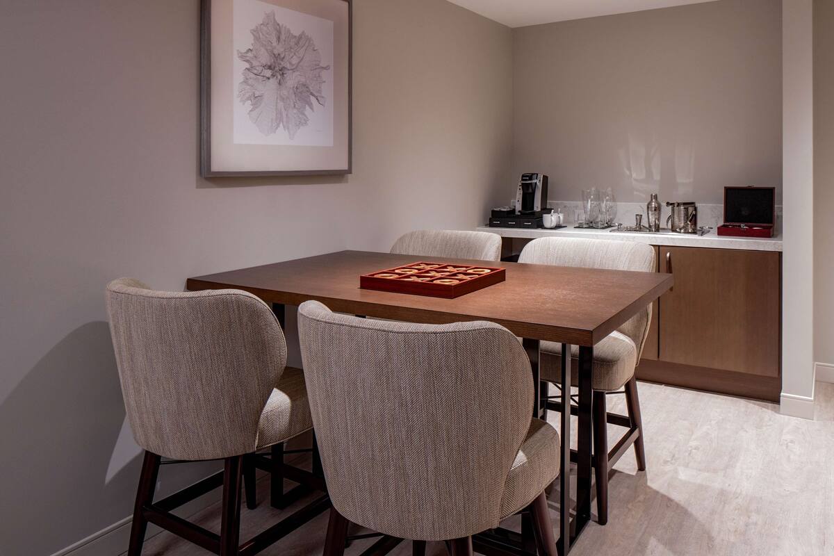 A dining area with a wooden table and four chairs, a framed artwork on the wall, and a kitchen area with appliances and glassware.