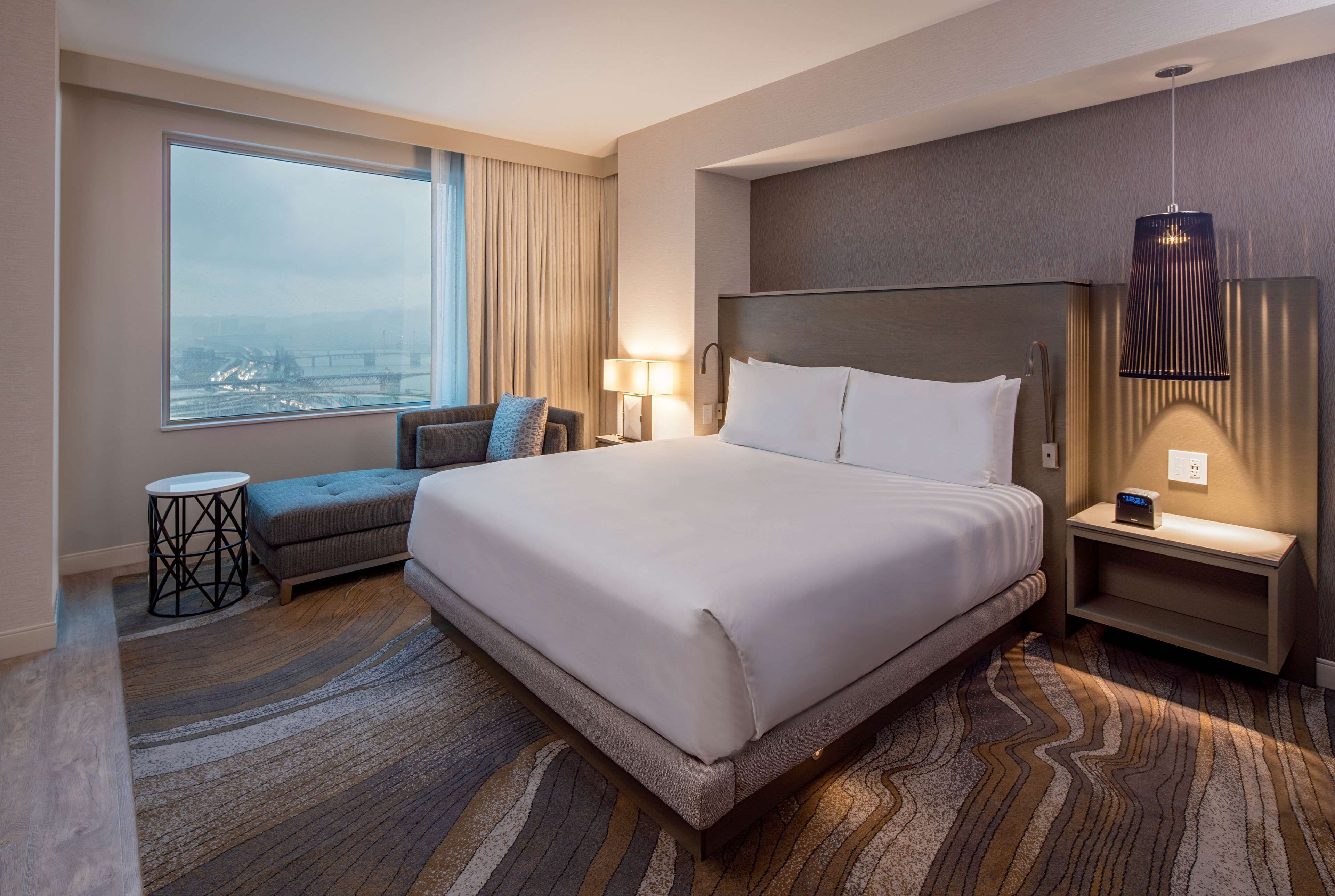 A modern hotel room with a large bed, a sofa, a small table, and a view of a bridge.