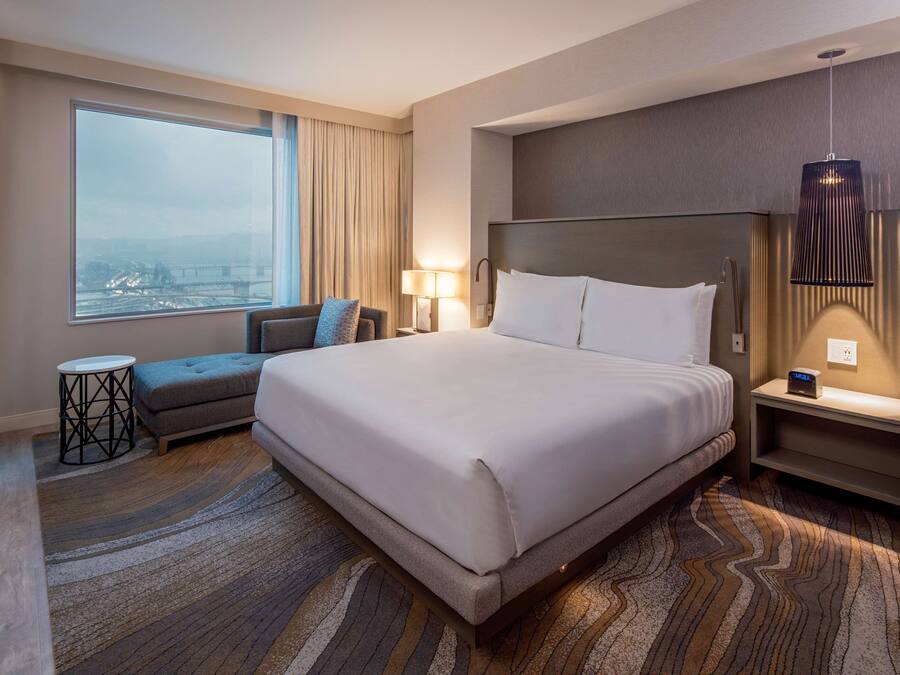 A modern hotel room with a large bed, a sofa, a small table, and a view of a bridge.