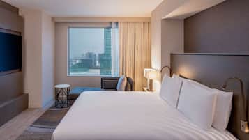 Quarto de hotel com uma cama grande, televisão na parede, janela com vista para a cidade, uma mesinha e uma luminária.