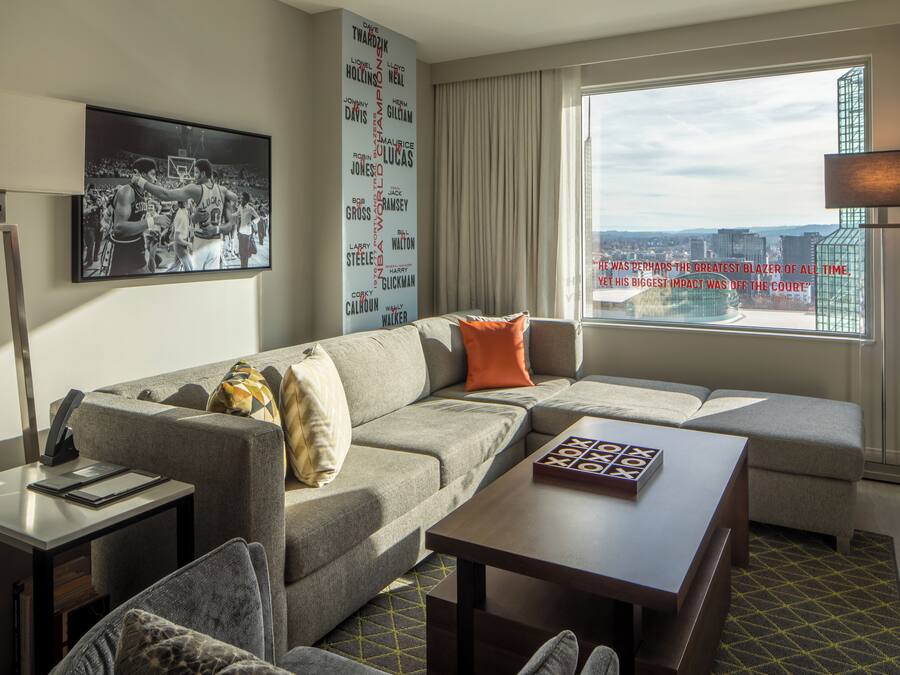 A modern hotel room with a sectional sofa, a small table, a desk with a lamp, and a large window offering a city view.