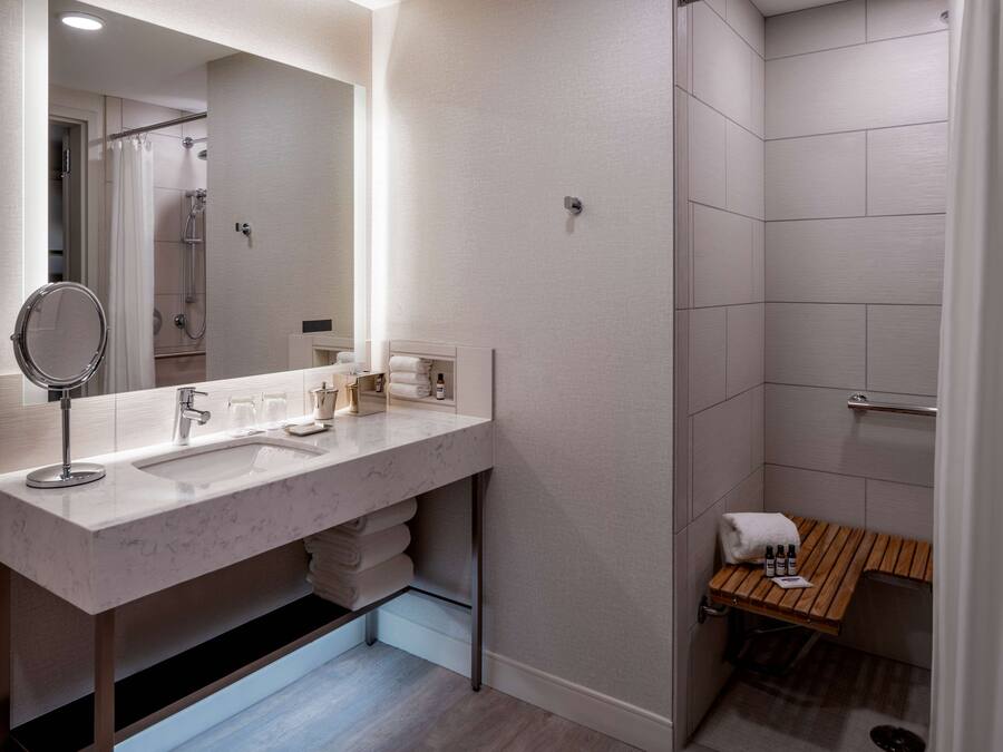 A modern bathroom with a large mirror, two sinks, a shower, and a wooden bench.