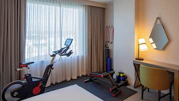 A hotel room with a bed, exercise equipment, a desk with a lamp, and a window with curtains.