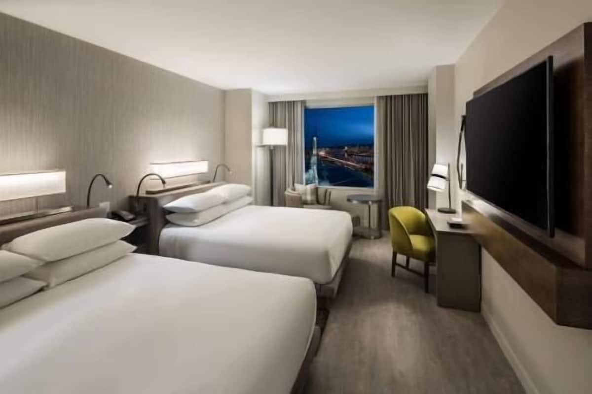 A hotel room with two beds, a desk, a chair, a TV, and a window with a view.
