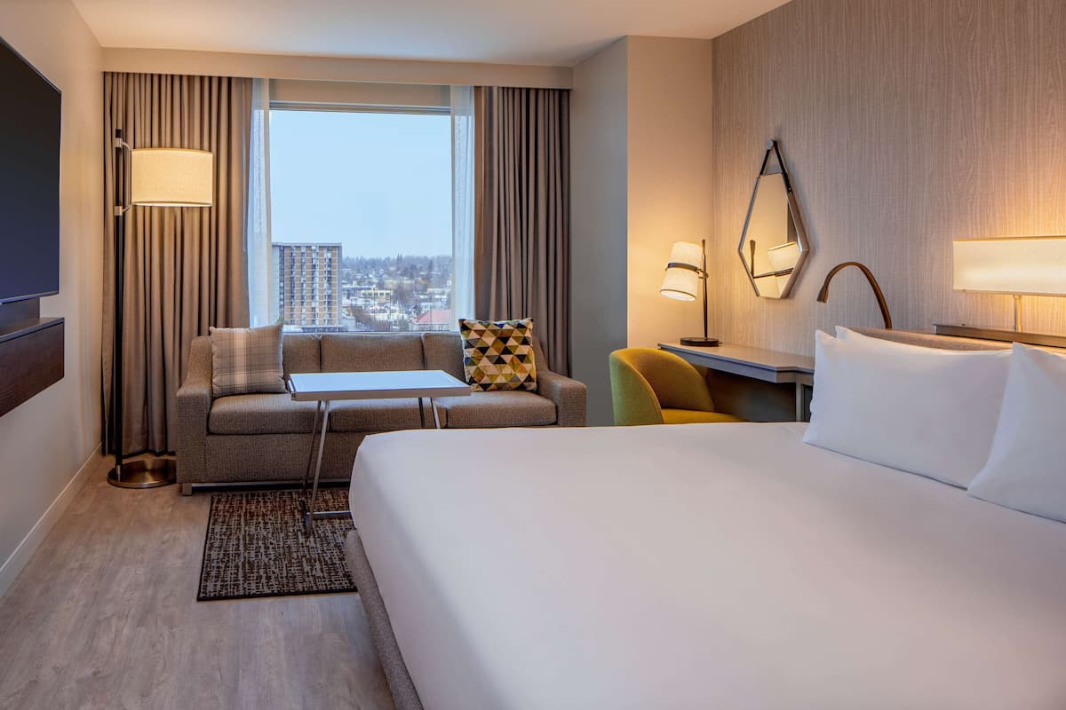 A hotel room with a large bed, a sofa, a small table, and a chair. There is a window with curtains, a lamp, and a mirror on the wall.