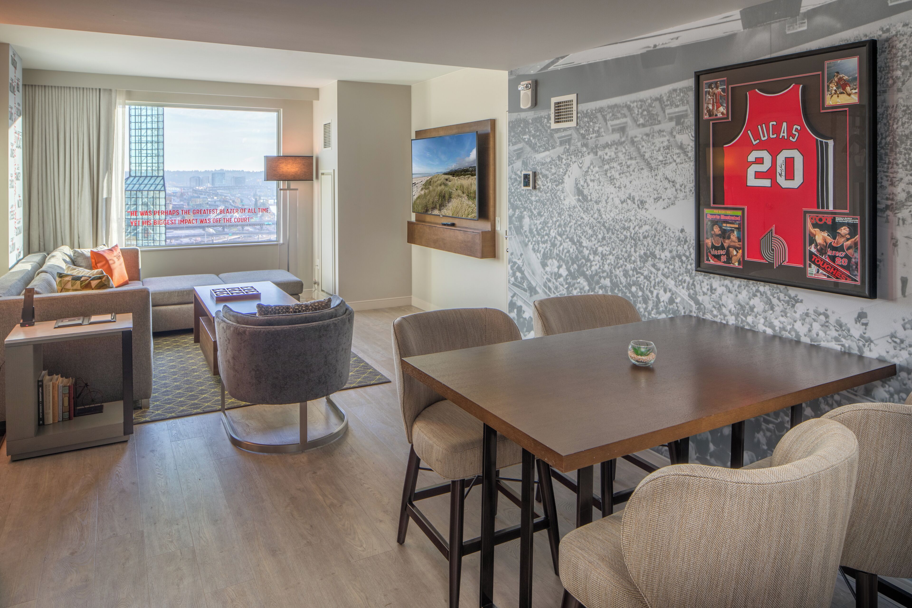 Una habitación de hotel moderna con un ventanal grande, un televisor de pantalla plana, una zona de comedor con mesa y sillas, y una pared decorada con recuerdos deportivos.