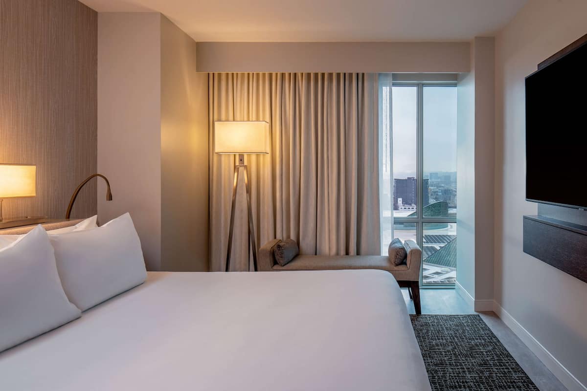 A hotel room with a large bed, a desk, a chair, a TV, a window with curtains, and a view of the city.