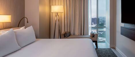 Ein Hotelzimmer mit einem großen Bett, einem Schreibtisch, einem Sessel, einem Fernseher, einem Fenster mit Vorhängen und Blick auf die Stadt.