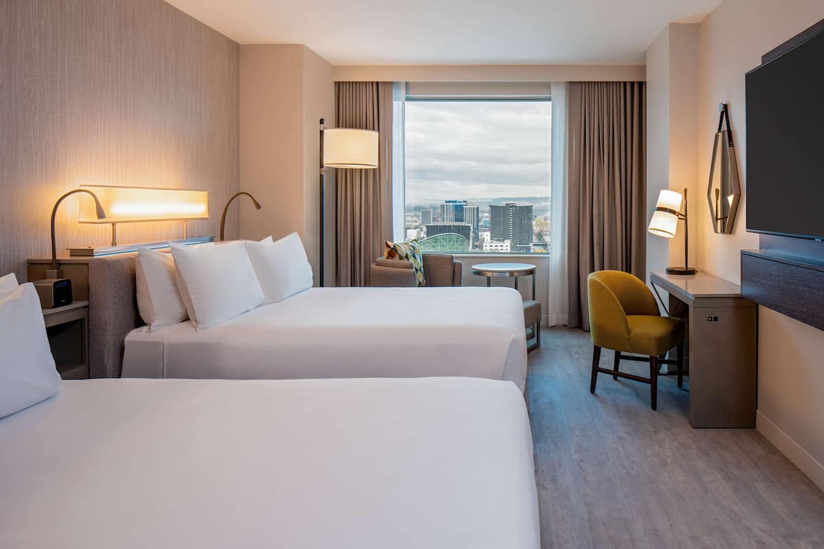 A hotel room with two beds, a desk, a chair, a TV, and a window with a city view.
