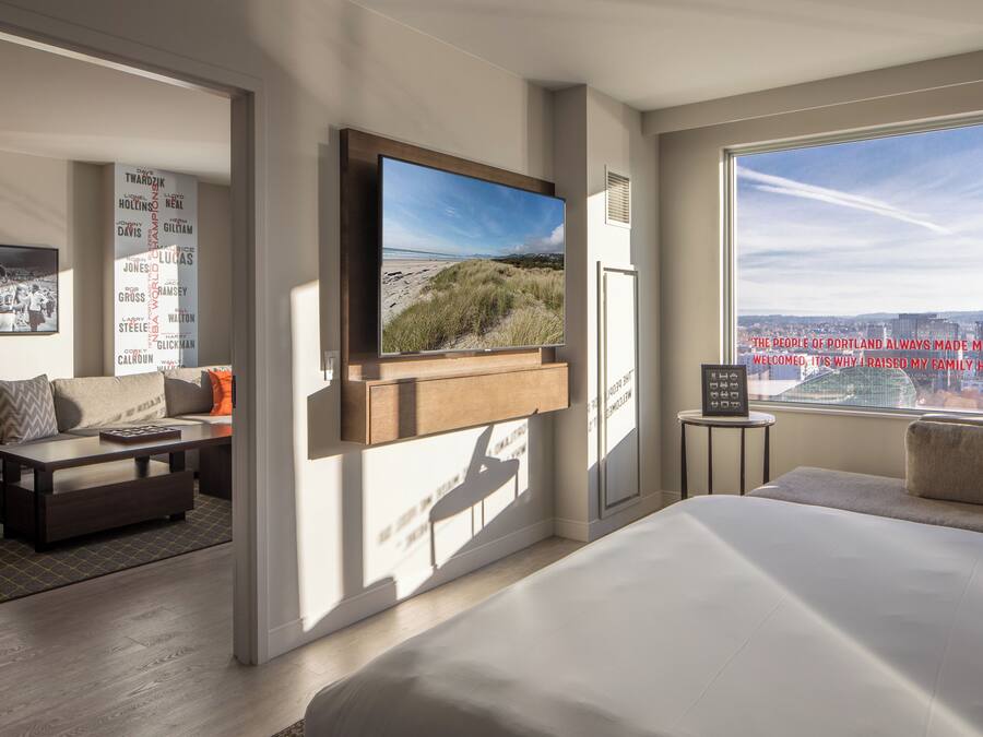A modern hotel room with a large TV mounted on the wall, a comfortable sofa, a bed, and a view of the city.