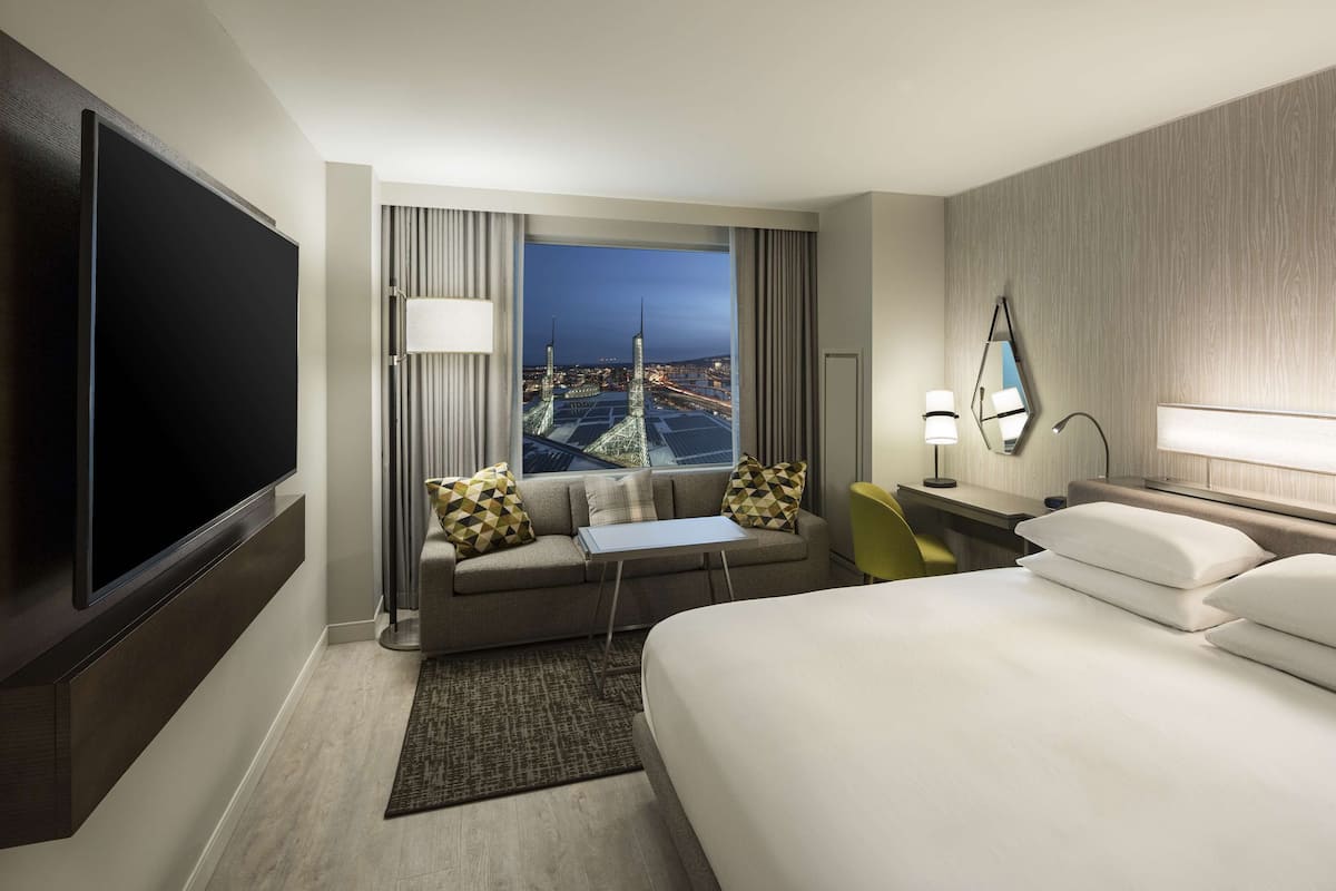 A hotel room with a bed, a sofa, a small table, a TV, and a view of a cityscape.