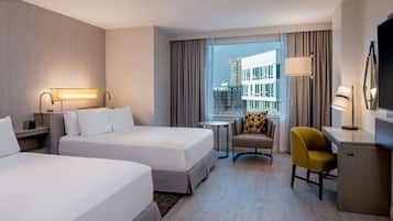 A hotel room with two beds, a desk, a chair, a TV, and a view of the city.