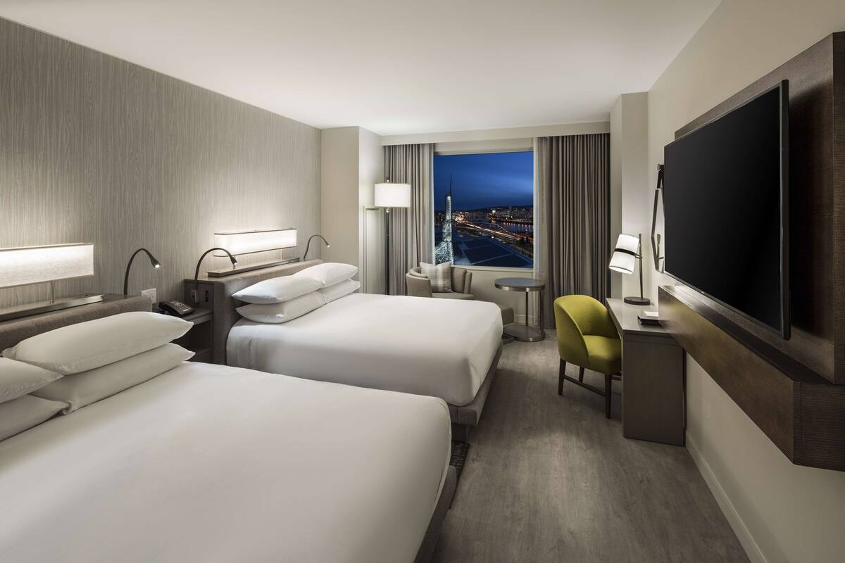 A hotel room with two beds, a desk, a chair, a TV, and a window with a city view.