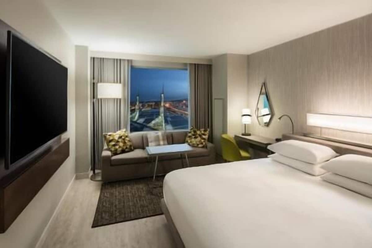 A hotel room with a bed, a sofa, a TV, a window with a city view, and a desk with a lamp.