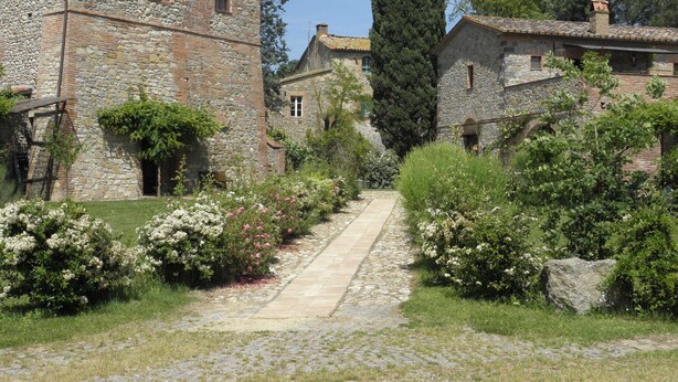 Un bâtiment en pierre, accessible par un chemin pavé, entouré d’une végétation luxuriante et de plantes en fleurs.