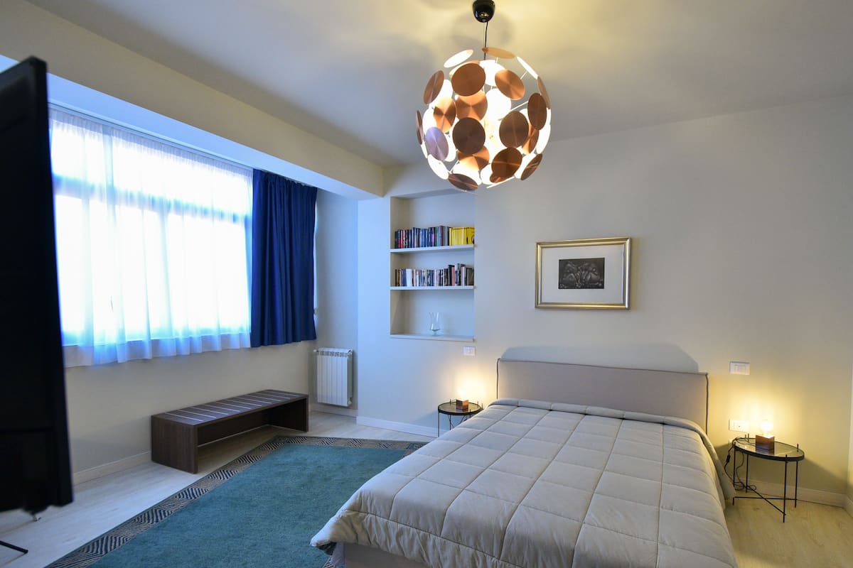 A bedroom with a large bed, a reading chair, a bookshelf, and a unique spherical light fixture.
