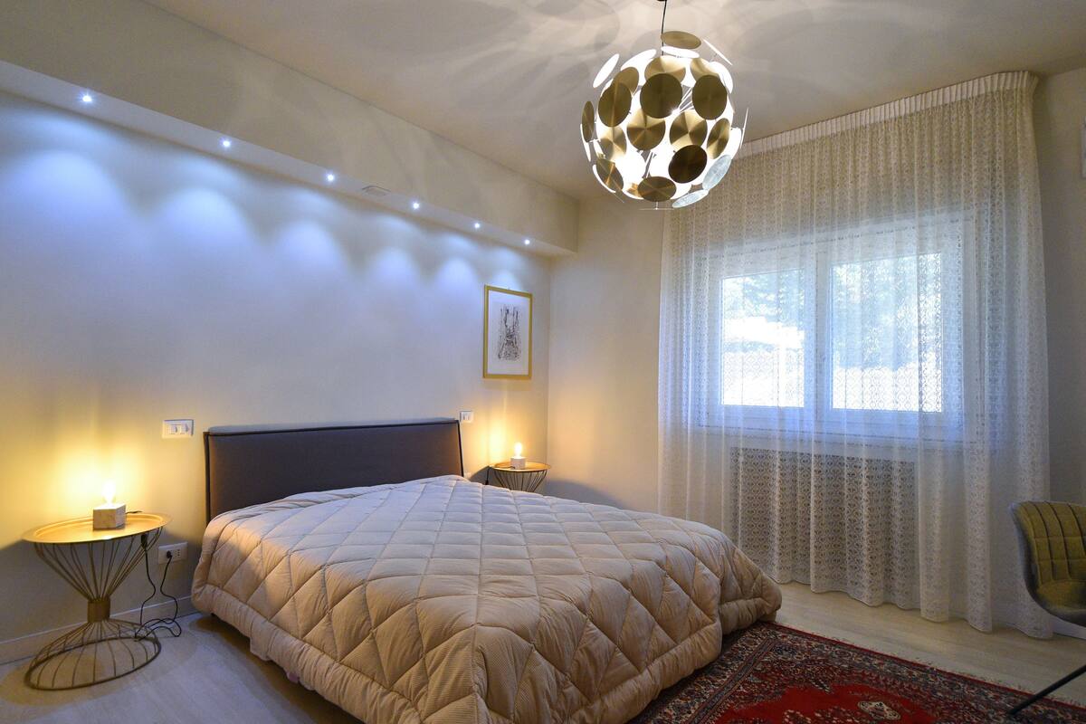 A bedroom with a large bed, bedside lamps, a decorative ceiling light, a window with sheer curtains, and a patterned rug.