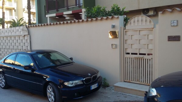 Un BMW azul oscuro estacionado frente a una casa color crema con entrada protegida por una reja y un balcón pequeño.