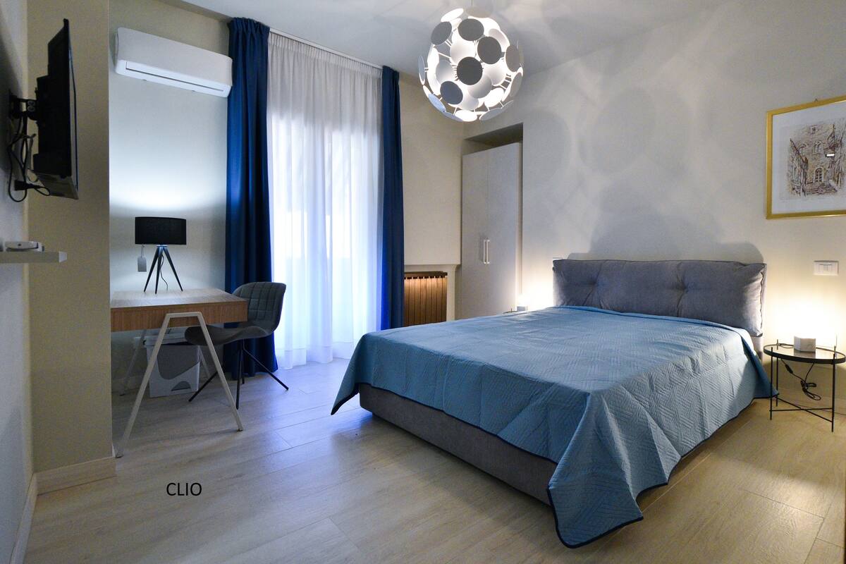 A modern hotel room with a large bed, a desk with a chair, a television, and a unique ceiling light.