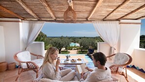 Two people sitting on wicker chairs on a tiled patio with a view of a pool and trees.