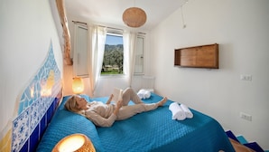 A room with a bed, a window with curtains, a hanging light fixture, and a wall-mounted shelf.