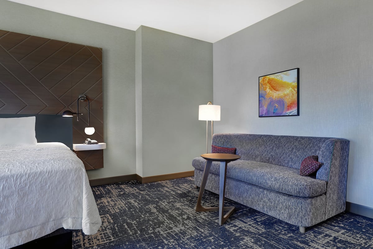 A hotel room with a bed, a sofa, a small table with a lamp, and a painting on the wall.
