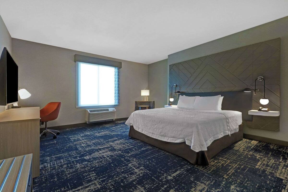 A hotel room with a large bed, a desk with a chair, a window with blinds, and a wall-mounted shelf with a lamp.