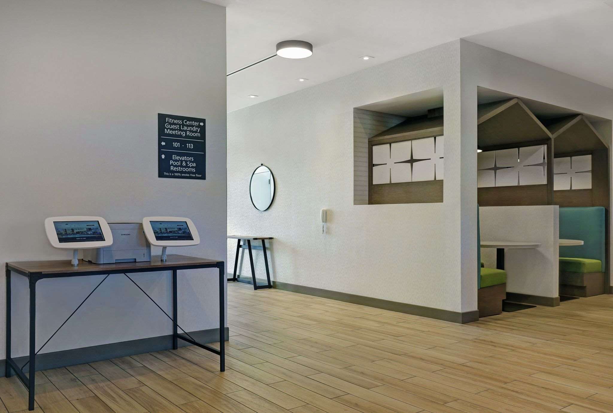 A hotel lobby with a reception desk, a sign indicating the fitness center, guest laundry, and meeting room, and a small table with a clock.