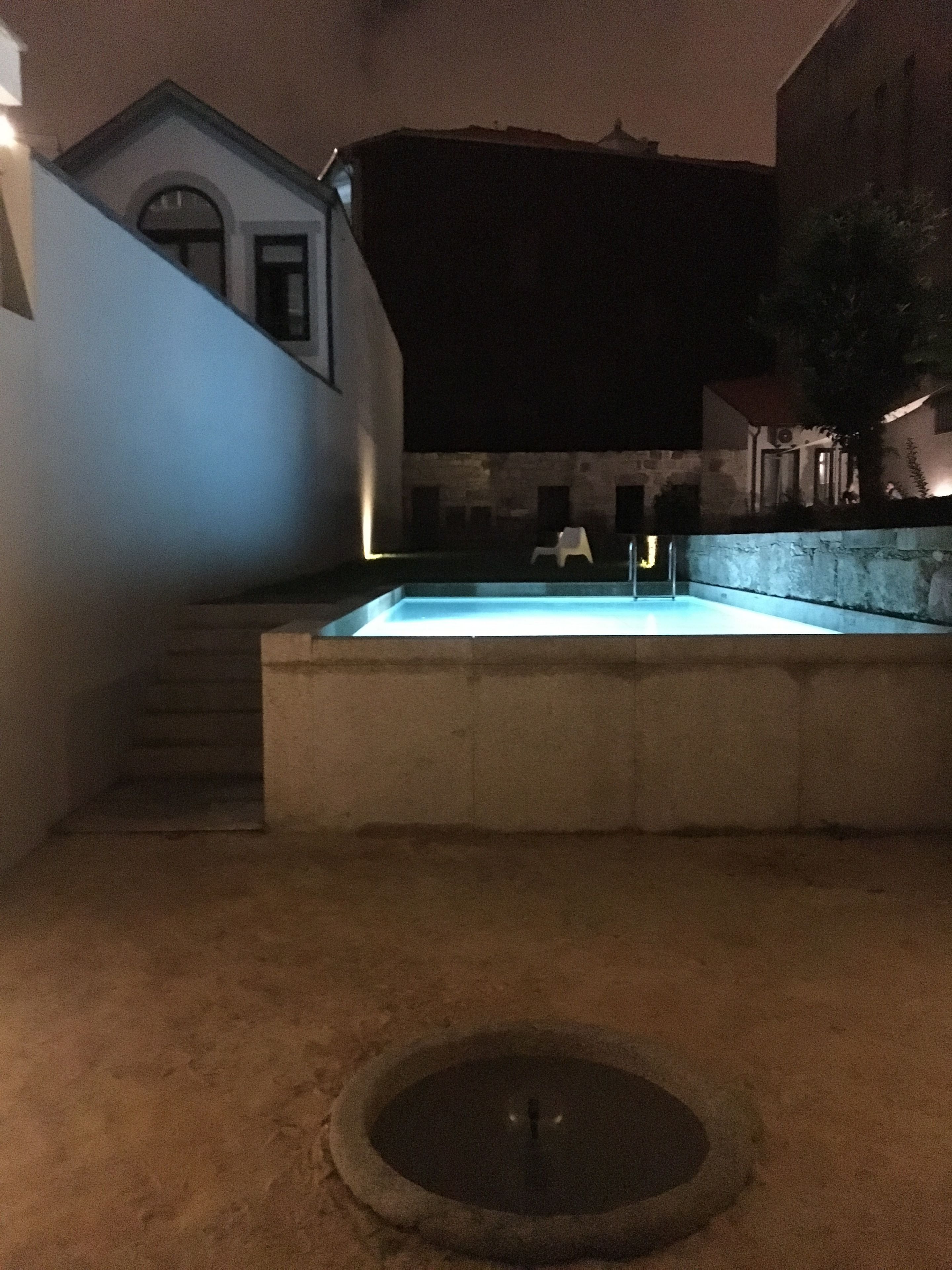 A nighttime scene of a swimming pool area with a stone border, a small fountain, and a white chair.